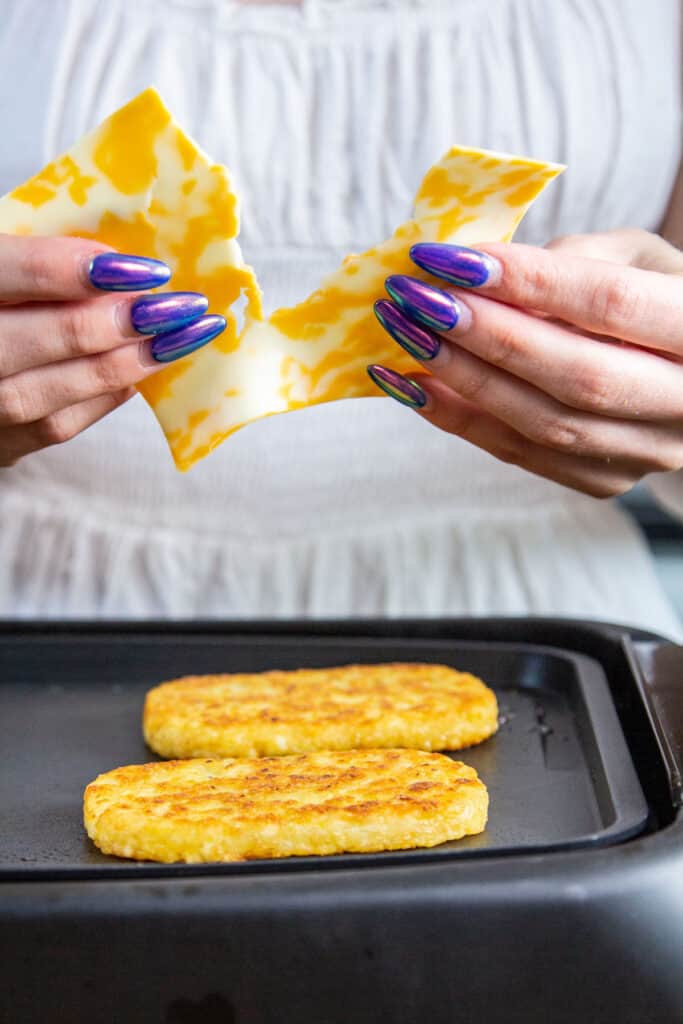 hand holding a slice of colby jack cheese over hash brown patties on a griddle